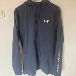 Blå vindjacka från Under Armour - Snygg vindjacka från Under Armour med huva och hel dragkedja. Lätt och smidig med långa ärmar och praktiska fickor.