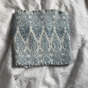 Blåvit bandeau tubtopp med mönster - Supersöt bandeau tubtopp i blått och vitt med ett vävt, nästan boho-inspirerat mönster. Toppen är elastisk och har en smockad struktur som gör att den sitter på plats. Perfekt till sommaren och lätt att matcha med jeans eller kjol.