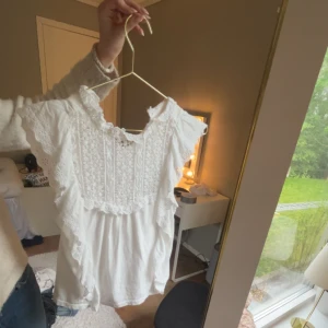 Vit blus med spets och volang - Supersöt vit blus med broderade spetsdetaljer och volanger längs ärmar och bröst. Blusen har korta ärmar och ett fint mönster framtill. Perfekt för dig som vill ha en fin blus till sommaren💘🌸