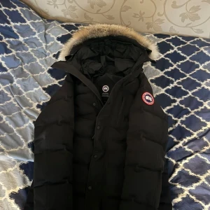 Svart dunjacka med pälskant från Canada Goose - Säljer en svart dunjacka från Canada Goose med huva och pälskant. Jackan har lång ärm, knappar och dragkedja framtill samt den klassiska loggan på ärmen. Perfekt för kalla dagar.
