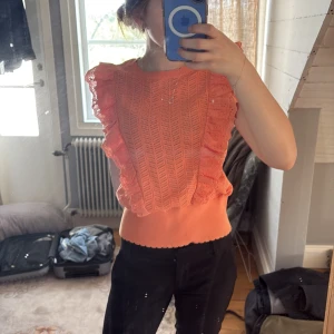 Korallfärgad ärmlös topp med volang - Superfin korallfärgad topp med breda volanger längs axlarna och ett snyggt hålmönster. Toppen är ärmlös och har en ribbad midja som ger en smickrande passform. Perfekt för att liva upp garderoben!