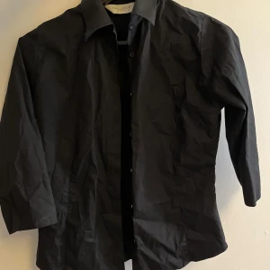 Svart skjorta från Russell Collection - Klassisk svart skjorta från Russell Collection med krage och knäppning framtill. Perfekt för en stilren look och enkel att matcha med olika outfits. Skjortan har lång ärm och är tillverkad i ett mjukt material.