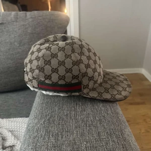 Beige keps från Gucci med GG-mönster - Säljer en beige keps från Gucci med klassiskt GG-monogram över hela kepsen och en grön-röd rand på sidan. Kepsen har böjd skärm och är tillverkad i ett tygmaterial med exklusiv känsla.