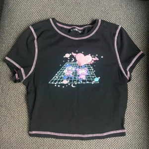 Svart croppad topp med rosa sömmar SHEIN - Svart croppad topp från SHEIN med kontrastsömmar i rosa och ett färgglatt tryck framtill med två tecknade figurer, hjärta med vingar och rymdtema. Toppen har korta ärmar och rund hals. Perfekt för dig som gillar kawaii och unik stil.
