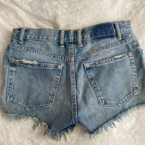 Blå jeansshorts med slitningar från Zara - Snygga blå jeansshorts från Zara med medelhög midja, slitna detaljer och fransig kant. Klassisk femficksmodell med knappar framtill. Perfekta för sommaren. 