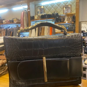 Svart handväska med kedjedetalj - Snygg svart handväska i croco-mönstrat skinnimitation med bred dekorativ rosett framtill och guldfärgad metallstav. Handtaget har en cool guldkedja som detalj. Perfekt för dig som vill sticka ut med en elegant och trendig accessoar.
