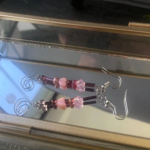 Vackra örhängen med rosa och lila pärlor - Unika handgjorda örhängen med mix av rosa och lila pärlor i olika former, inklusive hjärtformade och facetterade pärlor. Hängande design med silvriga krokar och dekorativa spiraler längst ner.