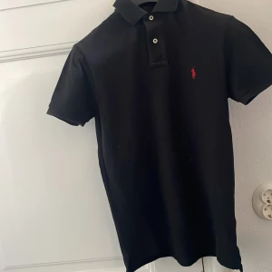 Svart pikétröja från Polo Ralph Lauren - Klassisk svart pikétröja från Polo Ralph Lauren med röd broderad logga på bröstet. Tröjan har korta ärmar, krage och två knappar framtill. Tillverkad i mjuk bomull som känns skön mot huden. Perfekt för en clean och stilren look.