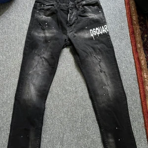 Svarta Dsquared2 jeans Squid Game Helt Ny! - Snygga svarta jeans från Dsquared2 med Squid Game-tag, vita DSQUAR-tryck på ena benet och coola slitningar samt färgstänk. Jeansen har smal passform och är tillverkade i bomull och polyester. Perfekta för dig som vill sticka ut med en edgy streetstil. Helt ny!! 