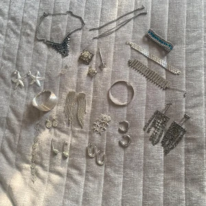 Stort paket med silvriga smycken - Säljer ett mixat paket med silvriga smycken: flera armband, halsband, en hårklämma, en brosch och örhängen. Här finns stjärnformade örhängen, glittriga statementörhängen, chunky armband, smala armband med blå stenar och flera olika hoops. Perfekt för dig som gillar att variera din stil och mixa accessoarer.