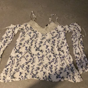 Vit blommig blus med spetsdetalj - Supersöt vit blus med blått blommönster och tunna axelband. Blusen har långa ärmar med knytband och v-ringning med spetsdetalj framtill. Perfekt för dig som gillar romantisk och feminin stil.