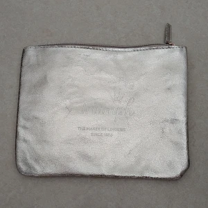 Silvrig kuvertväska från Triumph - Säljer en stilren silvrig kuvertväska från Triumph med dragkedja upptill. Väskan har en metallic finish och är fodrad med ljusrosa tyg på insidan. Perfekt för att bära småsaker eller använda som accessoar.