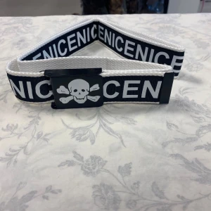 Svart och vitt bälte med dödskalle och text - Säljer ett svart och vitt bälte med texten 'NICENICENICE' runt om och ett spänne med dödskalle- och korsade ben-motiv. Bältet har en sportig och edgy stil, perfekt för att ge outfiten attityd. Helt ny 