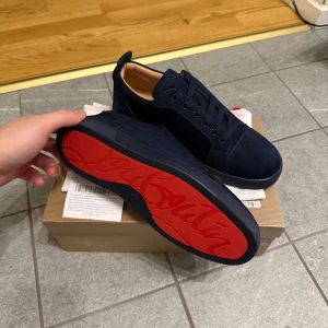 Mörkblå loubs i storlek 42 - Säljer ett par mörkblå sneakers från Louboutin i mocka med klassisk röd sula och snygg snörning. Skorna har en stilren design med rund tå och diskreta detaljer, perfekt för dig som vill sticka ut med lyxiga vibes.