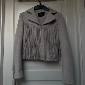 Beige mockajacka med fransar från Bik Bok - Säljer en beige mockajacka från Bik Bok med dragkedja och långa fransar både fram, bak och längs ärmarna. Jackan har klassisk krage och croppad passform. Perfekt statement-plagg för dig som vill sticka ut.