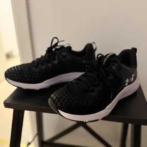 Svarta träningsskor från Under Armour - Säljer ett par svarta träningsskor från Under Armour med vit sula och snörning. Skorna har en sportig design med ventilerande mesh och logga på sidan. Perfekta för gym eller löpning. 