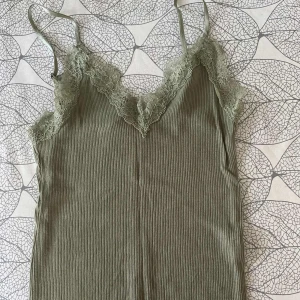 Olivgrön ribbad linnetopp med spets - Säljer en olivgrön ribbad linnetopp med tunna axelband och vacker spetsdetalj längs urringningen. Perfekt att styla till jeans eller kjol för en trendig look.