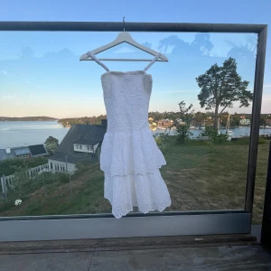 Vit somrig klänning med volang från RYVLS - Supersöt vit klänning från RYVLS med smock upptill och volangkjol. Axelbandsmodell och kort längd axelbanden är jätte lätta att ta av, perfekt för varma dagar. Klänningen har broderade detaljer och är tillverkad i bomull för en luftig känsla.