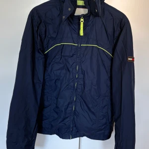 Blå vindjacka från Tommy Hilfiger Denim - Snygg blå vindjacka från Tommy Hilfiger Denim med gula detaljer och dragkedja. Jackan har lång ärm, hög krage och ett broderat logomärke på ärmen. Perfekt för svala dagar och enkel att matcha med olika outfits. I fin skick