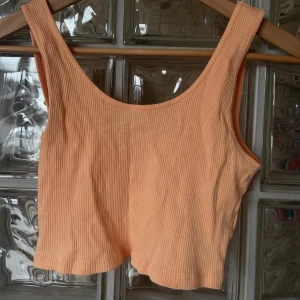 Persikofärgad ribbad linnetopp - En persikofärgad ribbad linnetopp med breda axelband och rundad halsringning. Toppen är croppad och har en mjuk, stretchig känsla som passar perfekt till sommaren.