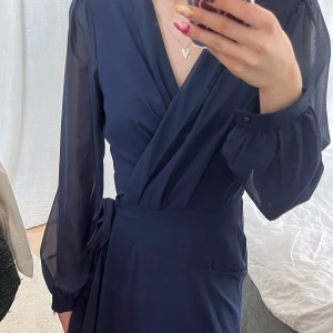 Mörkblå omlottklänning med långa ärmar - Säljer en elegant mörkblå långklänning med omlottmodell och v-ringning. Klänningen har långa, lätt transparenta ärmar med knappar vid ärmslut och ett knytband i midjan. Tyget faller snyggt och ger en flowy känsla. Perfekt för dig som vill ha en stilren och klassisk look.