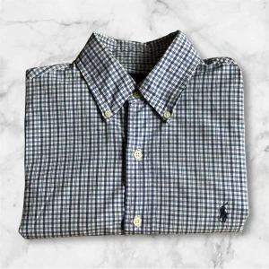 Blå Rutig skjorta från Ralph Lauren - Klassisk rutig skjorta från Ralph Lauren i blått och vitt. Skjortan har lång ärm, knappar framtill och ikonisk broderad logga på bröstet. Tillverkad i mjuk bomull och har en snygg, tidlös passform som funkar till många stilar. Tyvärr en liten fläck längst ner vid sista knappen men syns inte om man inte tittar noggrant