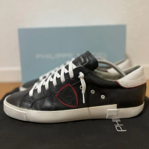 Svarta Philippe Model sneakers  - Snygga svarta sneakers från Philippe Model med vit sula och vita skosnören. Skorna har en vit hälkappa och röd detaljsöm runt logotypen på sidan. Tillverkade i skinn och har en klassisk låg profil som passar till många stilar. Storlek 44 passar också 43, bra skick. Liten defekt på högra toecap, fast inget som påverkar funktionaliteten eller helhetsintrycket.