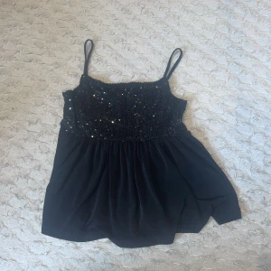 Svart paljettopp från Gina Tricot - Supersnygg svart topp från Gina Tricot med smala axelband och glittriga paljetter över bysten. Toppen har en luftig, lätt utställd passform och är perfekt för dig som vill sticka ut lite extra. Materialet känns mjukt och faller snyggt.