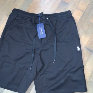 Svarta shorts från Polo Ralph Lauren - SELGER FRA NORGE SÅ ER NORSKE KRONER Svarta shorts från Polo Ralph Lauren med klassisk vit broderad logga på benet. Modellen har elastisk midja med snörning och sidfickor. Perfekta för en avslappnad och stilren look. Materialet känns mjukt och bekvämt mot huden.