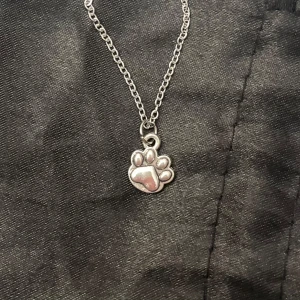 Tass halsband silver imitation  - Sött halsband med ett silverfärgat  tassavtryck. Berlocken är ca 0,7 x 0,6 cm. Kedjan är i silverimitation och justerbar 40-45 cm. 