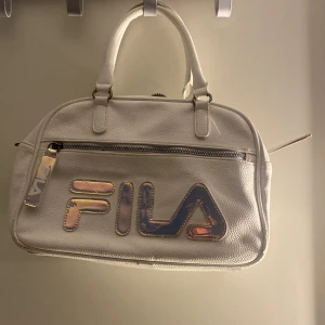 Vit axelväska från FILA - Säljer en vit axelväska från FILA med stort holografiskt logotyptryck på framsidan. Väskan har ett ytterfack med dragkedja och två handtag upptill. Perfekt för att bära dina viktigaste saker med stil.