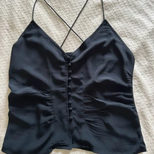 Svart linnetopp med knappar från Bik Bok - Snygg svart linnetopp från Bik Bok med smala axelband som korsas i ryggen. Toppen har dekorativa knappar framtill och en lätt v-ringad halsringning. Perfekt att styla till jeans eller kjol.