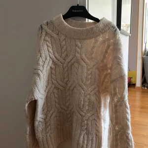 Vit stickad kabelmönstrad tröja från KappAhl - Mysig vit stickad tröja från KappAhl med grovt kabelstickat mönster och bred ribbad krage. Perfekt för kyliga dagar och enkel att matcha med jeans eller kjol.