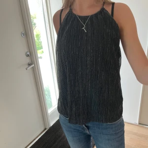 Svart glittrig halterneck  - Säljer en svart glittrig linnetopp från Divided med tunna axelband och rak passform. Toppen har ett diskret silvrigt skimmer i tyget och är perfekt för att lyfta en outfit. Supersnygg till både jeans och kjol!