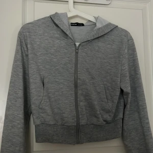Grå zip-hoodie från SHEIN - Tunn grå kofta med dragkedja från SHEIN. Modellen har huva, långa ärmar och ribbade muddar vid ärmslut och nederkant. Perfekt för en avslappnad stil och enkel att matcha med det mesta. Storlek S
