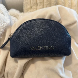 Mörkblå plånbok från Valentino - Säljer en stilren mörkblå plånbok från Valentino i skinn med silverfärgad dragkedja. Halvmåneformad design och präglad logga framtill. Perfekt för att hålla ordning på småsaker i väskan.
