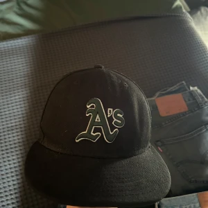 Svart keps Oakland A's från New Era - Svart keps från New Era med Oakland A's logga framtill och New Era-logga på sidan. Klassisk böjd skärm och broderade detaljer. Storlek 7 (55,8 cm). Perfekt för dig som gillar sportig stil.