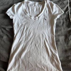Vit v-ringad t-shirt från Tommy Hilfiger - Vit t-shirt från Tommy Hilfiger med v-ringning och liten broderad logga på bröstet. Modellen är figurnära och har korta ärmar. Tillverkad i mjuk bomull som känns skön mot huden och perfekt för sommaren och när det blir varmt.😏
