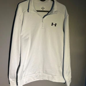 Vit träningströja med dragkedja från Under Armour - Säljer en vit långärmad träningströja från Under Armour med halv dragkedja och hög krage. Perfekt för sport eller vardag, med svart logga på bröstet. Skön passform och stilren design.