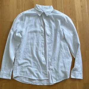 Vit randig skjorta från H&M, L - Vit skjorta från H&M i storlek L med tunna svarta ränder. Klassisk krage, långärmad och knäppning framtill. Regular fit-modell i lätt och luftig bomull, perfekt för en clean och stilren look. Inga defekter.