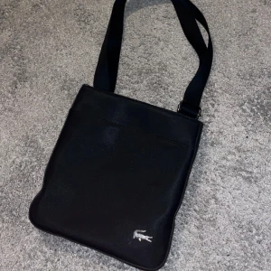 Svart axelväska från Lacoste - Snygg svart axelväska från Lacoste med klassisk krokodillogga i silver framtill. Väskan har justerbar rem, dragkedja upptill och ett innerfack med dragkedja. Perfekt storlek för det viktigaste och stilren design som funkar till allt.