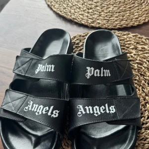 Palm Angels svarta sandaler i skinn - Svarta sandaler från Palm Angels med två breda remmar i skinn och vit logotyptext på ovansidan. Snygg och stilren design med bekväm fotbädd, perfekt för en avslappnad streetwear-look.