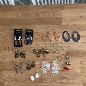 Stort paket med örhängen och halsband - Säljer ett paket med flera par örhängen i olika färger och former, bland annat guld, silver, svart och rött. Inkluderar både hängande och studs samt ett långt halsband i guldton med vita detaljer. Perfekt för dig som vill variera din stil.