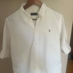 Vit skjorta från Polo Ralph Lauren - Klassisk vit skjorta från Polo Ralph Lauren med broderad logga på bröstet. Skjortan har knappar framtill, krage och korta ärmar. Perfekt för en clean och stilren look. Tillverkad i mjuk bomull för maximal komfort.