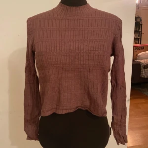 Brun/rosa ribbad långärmad tröja - Säljer en dauve-rosa ribbad långärmad tröja med hög hals. Tröjan har ett snyggt strukturerat mönster och är croppad i modellen. Perfekt för dig som gillar en enkel men trendig stil. Passar till både jeans och kjol.