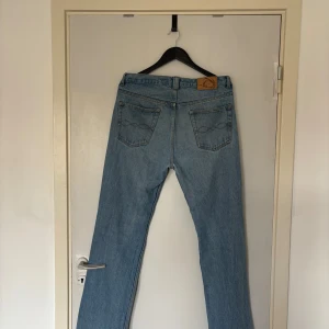  ljusblå jeans W34 L32 - Klassiska ljusblå denim. Rak passform med fem fickor och knappgylf. Tillverkade i 100% bomull för en autentisk känsla. Perfekta för dig som gillar tidlös stil och vill ha ett par jeans som funkar till allt.