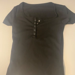 Svart ribbad topp med knappar - En svart ribbad topp med korta ärmar och snygg knappdetalj framtill. Toppen har en rundad halsringning och sitter figurnära. Perfekt att matcha med jeans eller kjol.🤗
