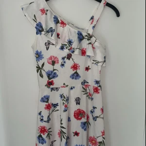 Blommig offshoulder sommarklänning - Superfin kortklänning med blommigt mönster i blått och rött på vit botten. Klänningen har en axelbandslös design med volangdetalj och en axelrem. Perfekt för varma dagar och ger en fräsch och ungdomlig look.