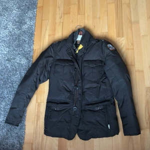 Svart Parajumpers dunjacka M - Snygg svart dunjacka från Parajumpers med klassisk patch på ärmen och cool gul detalj vid kragen. Jackan har flera fickor med både dragkedja och knappar, samt både dragkedja och knäppning framtill. Perfekt för kalla dagar och riktigt stilren look.