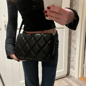 Svart axelväska - Säljer en svart axelväska med quiltad design och justerbar rem. Väskan har en stilren, rektangulär form och stängs med lock. Perfekt för dig som gillar en klassisk men trendig look. Materialet är skinnimitation och detaljerna är i guld. Kontakta mig ifall ni är intresserade eller om ni har frågor/fler bilder❣️❣️